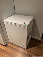 White Insignia chest freezer positioned in a corner against a wall on a wood floor.