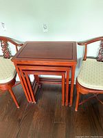 Frontal view of three wood nesting tables stacked tightly between two chairs on dark wood floor, showing the reddish-brown finish and rectangular design.