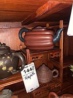 Accordion-ribbed brown clay teapot on top shelf of carved wood stand with part of dark brown floral teapot visible