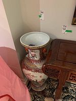 Full view of the tall porcelain vase placed on a carpet next to wooden furniture showing the wide funnel shaped rim and detailed hand-painted design including flowers and ornate borders.