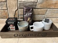 Coffee set displayed on wooden tray labeled 'Original' in front of stone wall. Includes 4 different ceramic mugs, unopened Starbucks Italian Roast coffee beans, black box labeled Bonus Ceramic Mug, and polka dot cloth inside one mug.