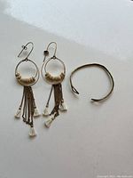 Photo showing pair of dangle earrings with wooden beads and hanging metal chains with white tassels alongside a brass cuff bracelet on a white surface.