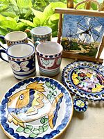 Photo of all lot items displaying 4 hand-painted mugs, 2 decorative plates, a framed tile painting, a ceramic knob, and a decorative tile against a garden background