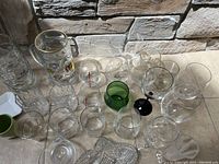 Various glassware items arranged on a counter including branded beer steins, multiple clear glass beer mugs and drinking glasses, a green small glass, and a black stemmed glass.