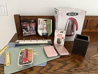 Photo of lot showing Bella electric kettle in box, brown leather-look container, pink sponge pads, candleholder kit, meat thermometer, and metal hooks.