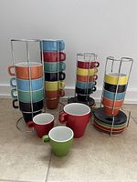 Photo of two vertical metal holders containing colorful stackable coffee mugs and espresso cups, with matching saucers at the base, arranged on a tile floor.