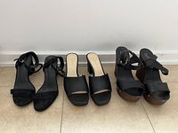 Side-by-side view of four pairs of black high heel shoes showing ankle straps, open toes, and various heel styles.