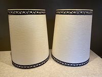 Two oversized tapered fabric lampshades with white textured fabric and black velvet decorative trim on top and bottom edges against a dark background.