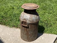 Milk can placed on pavement against green grass background showing full body and lid.