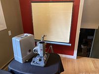 Photo shows the DeJur Model 1000-B 8MM film projector on a table with its original hard carry case closed beside it, and projection screen stretched open on a tripod stand behind them.