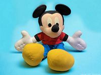 Front-facing image of the large plush Mickey Mouse wearing a red shirt, blue pants, yellow shoes, white gloves, sitting upright