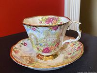 Front angled view of teacup and saucer showing floral garden and cottage scene decor with gold trim on cup handle and foot, and scalloped edges on both pieces.
