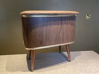 Side view of the hamper showing wood grain laminate exterior, cream trim, and tapered wooden legs with brass tips.