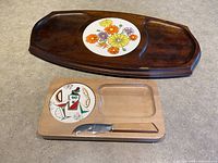 Photo showing both cheese boards, the larger dark wood one with floral ceramic inset on top, and smaller light wood one with colorful ceramic inset and cheese knife in carved slot