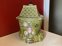 Front angle photo of ceramic cookie jar shaped like a shoe house with figures of an old woman and children at the door and window. The jar is olive green with detailed roof and windows.