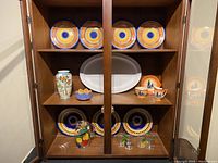 Closed walnut china cabinet containing decorative plates, vase, bowls, pitcher, and glasses on three shelves