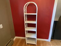 Front view of the white rattan wicker shelf against a red wall and wooden floor, showing five shelves and curved top.