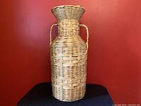 Full front view of tall multipurpose woven vessel made of wicker and rattan against a red wall background placed on black surface.