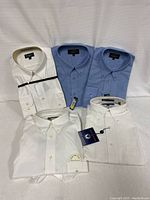 Five men’s long sleeve dress shirts neatly folded, 2 white, 2 light blue, and 1 white with striped collar, all button-up.
