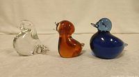Three glass duck paperweights arranged side by side, showing clear, amber, and blue colors and their relative sizes.