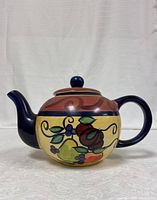 Side profile of hand painted ceramic teapot showing fruit design and blue handle and spout