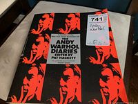 Front cover of The Andy Warhol Diaries paperback book showing red and black design with repeated Andy Warhol face, and text including editor and bestseller information.