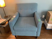 Front view of a blue upholstered chair with button tufted backrest and wooden legs beside a lamp and glass side table.