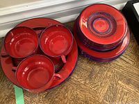 Photo showing stacked plates and three handled soup bowls in red with blue detailing on a wooden surface.