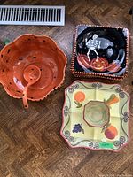 Large orange pumpkin-shaped ceramic bowl with attached dip bowl and ladle inside, black square plate with skeleton and pumpkin design, and harvest-themed serving platter with fruit and vegetables on a wood floor.