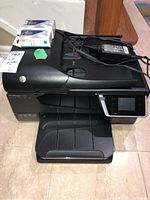 Front and top side view of HP Officejet 6600 with power adapter and two ink cartridges on top.