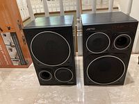 Pair of Jamo Compact 90 speakers side by side on floor showing front driver and port configuration, wood veneer sides.