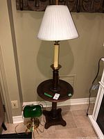 Photo showing the full view of the wooden table lamp with round side table and white pleated lampshade next to a green glass bankers desk lamp on the floor.