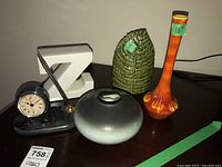 Photo of assorted decorative items including an orange ribbed vase, green leaf-patterned ceramic vase, matte gray-green rounded vase, and a Reflections desk clock with pen set.