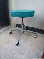 Side angled view of the vintage stool showing teal vinyl seat, metal adjustable screw, and caster wheels on metal base.