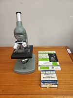 Front view of vintage Orion microscope with stage, three objective lenses, and eyepiece, also showing instruction manual and microscope slides box on table.