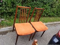 Pair of vintage teak chairs with tall slatted backs and orange cushions, placed outside on pavement with green hedge background.