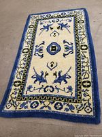 Full view of the rectangular vintage wool rug on a carpeted floor showing intricate design in blue, cream, and green colors with a distinct border and central motifs.