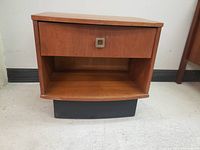 Front view of vintage teak night table showing drawer and open shelf with black base.
