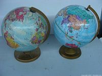 Two colorful political world globes on bronze-colored metal stands with meridian rings, viewed from front-left angle showing Pacific Ocean and Asia.