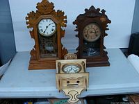 Three antique mantel clocks shown on a white table, varying in wood tone and height, untested condition.