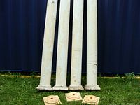 Four white pillars with peeling paint standing vertically on grass with four square bases placed in front.