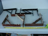 Photo shows six steel brackets laid out on a white surface. Four brackets are larger and reddish-brown; two brackets are smaller and black.