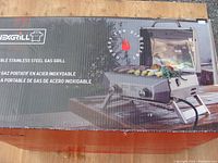 Photo of unopened box showing the NEXGRILL portable stainless steel gas grill with lid and side shelves, new packaging condition.