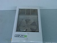 Front view of white wall-mounted electric heater with ventilation grill and fan behind it. Label with 'HEATER 220V TESTED DEC/17' is visible.