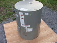 Full view of cylindrical grey electric water heater with access panels, warnings and labels. Positioned on wooden base outside.