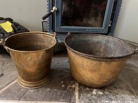 Two brass buckets side by side on stone fireplace hearth. One bucket is taller and narrower, the other larger and wider with two handles.