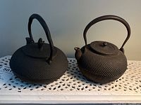 Photo of two cast iron teapots side by side on a white perforated surface. Both have textured surfaces, large curved handles, and lids. One has a scalloped handle and pinecone lid finial. The other has a round handle and knob lid finial.