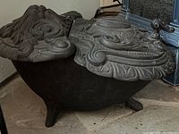 Side-angle photo of the black cast iron coal scuttle, showing the curved body and detailed embossed lid with a central rounded motif and intricate surrounding foliage and pattern work.