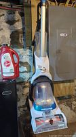 Full view of Hoover Power Dash Pet rug shampooer with the detergent bottle beside it in a basement setting.