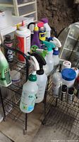Photo showing multiple household chemical spray bottles, including odor eliminator and WD-40, arranged on one wire metal shelf on wheels.